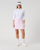 A model stands against a light gray studio background wearing a white long-sleeve collared golf shirt, a light pink mini skirt, white socks, white shoes, a white visor, and white gloves.