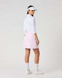 A woman modeling a light pink athletic skirt, paired with a white long-sleeve top, white visor, white socks, and white sneakers, shown from a side-back angle against a light gray studio background.