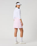 A woman modeling a light pink athletic skirt, paired with a white long-sleeve top, white visor, white socks, and white sneakers, shown from a side-back angle against a light gray studio background.