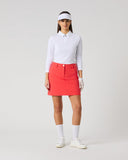 A model stands against a light gray studio background wearing a white long-sleeve collared polo shirt, a bright coral-red mini skirt, white socks, white shoes, and a white visor.