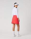 A woman stands in profile wearing a white long-sleeve top, a coral-red pleated athletic skirt, white socks, and a white visor against a light gray studio background.