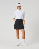 A woman modeling a white long-sleeve polo shirt, black golf skirt, white visor, white gloves, white socks, and white golf shoes against a light gray studio background.
