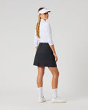 A woman modeling a white long-sleeve golf shirt, black pleated golf skirt, white visor, white socks, and white sneakers, shown from a side-back angle against a light gray studio background.