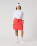 A woman modeling a white long-sleeve polo shirt and a bright coral-red A-line skirt, styled with a white visor, white gloves, white socks, and white sneakers against a light gray studio background.