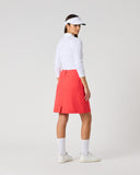 A woman stands in profile wearing a white long-sleeve top, a coral-red skirt, white socks, and white sneakers against a plain light gray studio background.