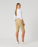 A woman stands in profile wearing beige knee-length shorts, a white long-sleeve top, and white sneakers against a light gray studio background.