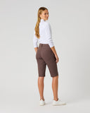 A woman stands in profile wearing brown fitted shorts, a white long-sleeve top, and white sneakers against a plain light gray studio background.