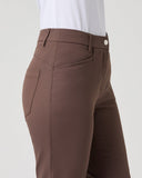 A close-up side view of a person wearing brown tailored trousers with a white top, shown against a plain light gray studio background.