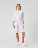 A model stands against a light gray studio background wearing a white long-sleeve collared top, light pink fitted shorts, white gloves, and white sneakers.