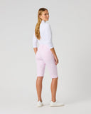 A woman stands in profile wearing light pink cropped shorts, a white long-sleeve top, and white sneakers against a plain light gray studio background.