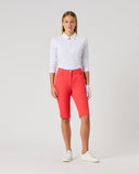 A woman modeling a white long-sleeve polo shirt and bright coral-red golf shorts, paired with white golf gloves and white sneakers, against a light gray studio background.