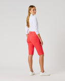 A woman standing in profile wearing bright coral-red cropped shorts, a white long-sleeve top, and white sneakers against a light gray studio background.