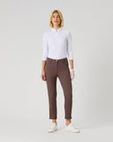 A woman modeling slim-fit brown trousers with a white long-sleeve polo shirt and white sneakers, standing against a light gray studio background.