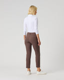 A woman is shown from behind wearing brown slim-fit trousers, a white long-sleeve mock neck top, and white sneakers, standing against a light gray studio background.