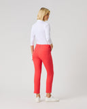 A woman with blonde hair is shown from behind wearing bright coral-red slim-fit pants, a white long-sleeve top, and white sneakers, standing against a light gray studio background.