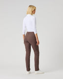 A woman with blonde hair is shown from the back wearing a white long-sleeve mock neck top, brown slim-fit trousers, and white sneakers, standing against a light gray studio background.