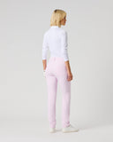 A woman with blonde hair is shown from behind wearing light pink slim-fit trousers, a white long-sleeve mock-neck top, and white sneakers, standing against a plain light gray studio background.