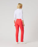 A woman standing with her back to the camera, wearing a white long-sleeve mock-neck top and bright coral-red slim-fit pants with white sneakers, against a light gray studio background.
