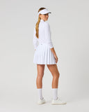 A woman modeling a white long-sleeve athletic top and matching pleated tennis skirt, paired with white socks, white sneakers, and a white visor, photographed against a seamless light gray studio background.