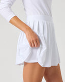 A close-up of a model wearing a white athletic tennis skirt and matching white long-sleeve top against a light gray studio background.