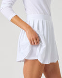 A close-up of a model wearing a white athletic tennis skirt and matching white long-sleeve top against a light gray studio background.