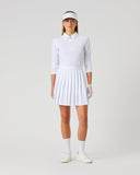 A woman modeling a white long-sleeve polo shirt and pleated tennis skirt set, complete with a white visor, white gloves, white socks, and white sneakers, against a solid light gray studio background.