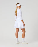 A woman modeling a white long-sleeve tennis dress with a pleated skirt, paired with a white visor, white socks, and white sneakers, photographed against a seamless light gray studio background.