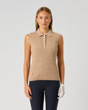 A woman stands against a light gray studio background wearing a sleeveless beige knit polo top with a collar and partial placket, paired with dark navy pants and a white golf glove.