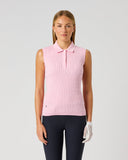 A woman stands against a light gray studio background wearing a sleeveless pink knit polo top, dark navy pants, and a white golf glove.
