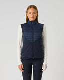 A woman modeling a navy blue sleeveless zip-front vest over a white long-sleeve top and dark leggings, standing against a light gray studio background.