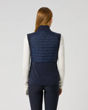A woman viewed from the back wearing a navy blue quilted puffer vest over a white long-sleeve top and dark navy leggings, against a plain light gray studio background.