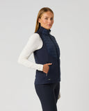 A woman modeling a navy blue quilted sleeveless vest over a white long-sleeve top and dark leggings, standing against a light gray studio background.