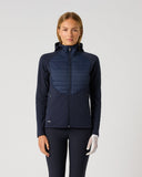 A woman stands against a light gray studio background wearing a dark navy hooded jacket with a quilted chest panel and matching dark navy leggings.