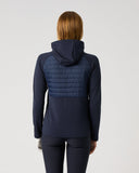 A woman viewed from the back wearing a navy hooded jacket with a quilted padded upper back panel, paired with dark leggings, against a plain light gray studio background.