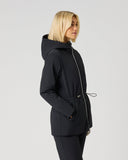 A woman with blonde hair wearing a black hooded jacket with a drawstring waist and black pants, standing in profile against a light gray studio background.