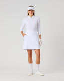 A woman modeling a white long-sleeve collared polo shirt dress with a matching skirt, white visor, white gloves, white socks, and white sneakers against a light gray studio background.