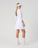 A woman modeling a white long-sleeve tennis dress, white visor, white socks, and white sneakers, standing in profile against a light gray studio background.