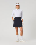 A female model stands against a light gray studio background wearing a white long-sleeve collared golf shirt, a dark navy skort, a white visor, white gloves, white socks, and white golf shoes.
