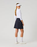A woman modeling a dark navy golf skirt, paired with a white long-sleeve top, white visor, white socks, and white golf shoes, photographed against a seamless light gray studio background.