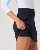 A close-up of a model wearing a dark navy wrap-style mini skirt with a white long-sleeve top, shown from the waist down against a light gray studio background.
