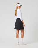 A woman stands in profile wearing a white long-sleeve athletic top, a black skirt, white socks, white sneakers, and a white visor against a plain light gray studio background.