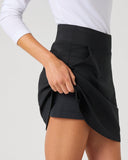 A close-up of a model wearing a black wrap-style athletic skirt, pulling the side fabric to show the layered shorts underneath. The model is also wearing a white long-sleeve top, set against a plain light gray studio background.