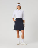 A woman modeling a white long-sleeve collared polo shirt and a dark navy blue skirt, styled with a white visor, white gloves, white socks, and white sneakers against a light gray studio background.