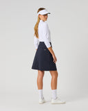 A woman modeling a dark navy blue athletic skirt, paired with a white long-sleeve top, white visor, white socks, and white sneakers, standing against a light gray studio background.