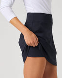 A close-up of a model wearing a dark navy wrap-style mini skirt with a white long-sleeve top, shown from the waist down against a light gray studio background.