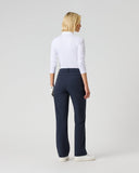 A woman stands with her back to the camera wearing a fitted white long-sleeve top, dark navy trousers, and white sneakers against a plain light gray studio background.