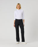 A woman stands against a light gray studio background wearing a white long-sleeve polo-style top, dark charcoal pants, and white sneakers.