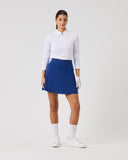 Woman wearing a white long-sleeve top, blue skirt, and white shoes on a light gray background