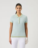 Woman wearing a light green checkered polo shirt and white pants on a plain background