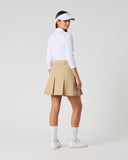 A woman stands in profile wearing a white long-sleeve top, a beige pleated tennis skirt, white socks, and a white visor against a light gray studio background.